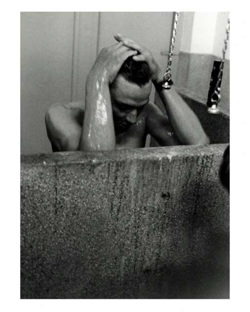 Black and white portrait of an adult male sitting in a bathtub, holding his head, photographed indoors with a close-up camera angle.