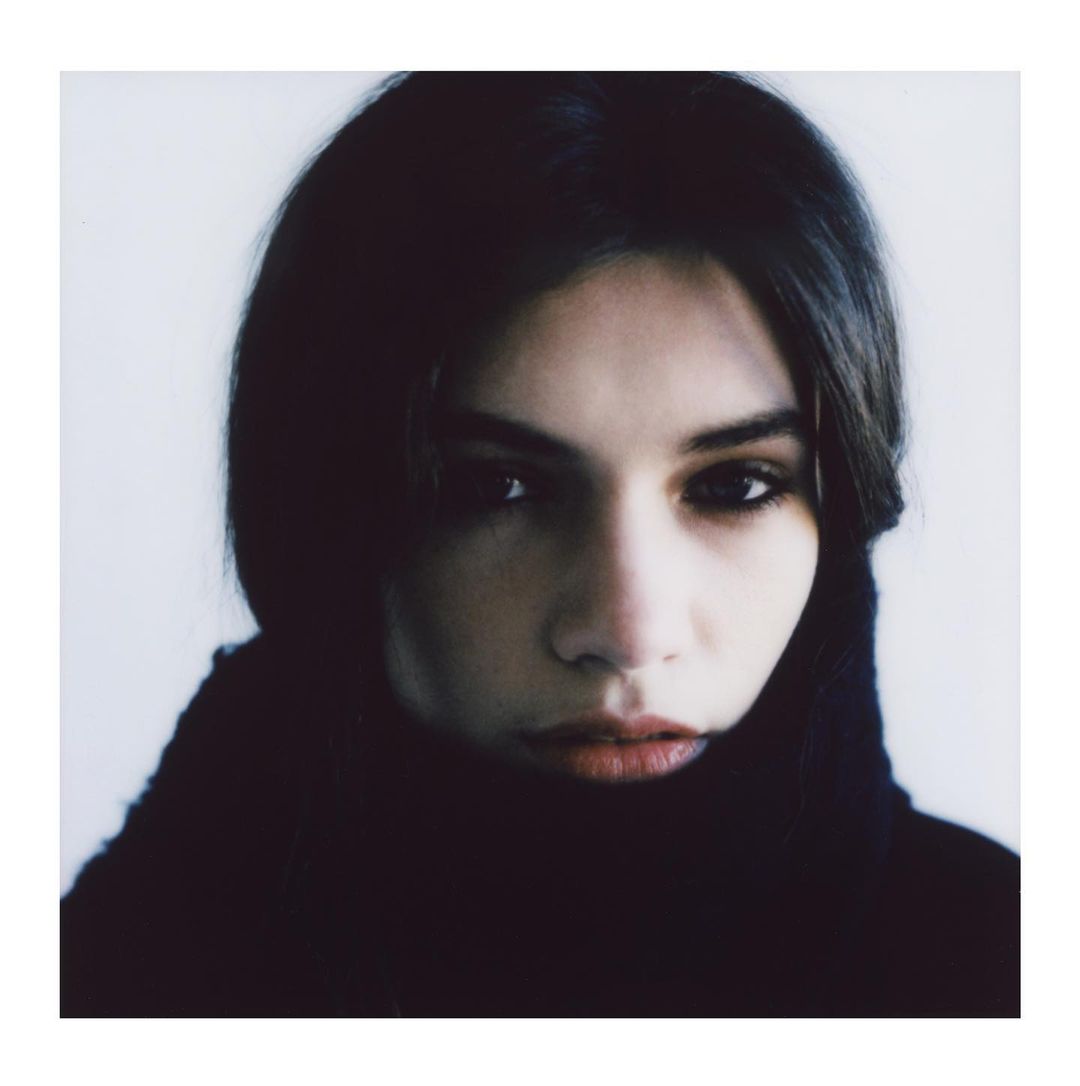 Close-up portrait of an adult woman with dark hair and eyes, wearing a dark turtleneck sweater against a muted grey-white backdrop.
