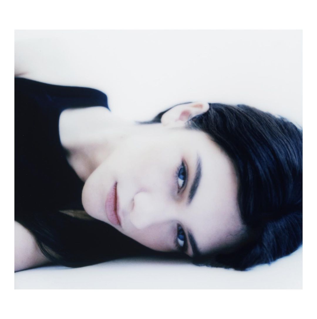 Close-up portrait of a young woman with dark hair and blue eyes, lying down in a light-filled, minimalist setting, focusing on beauty.