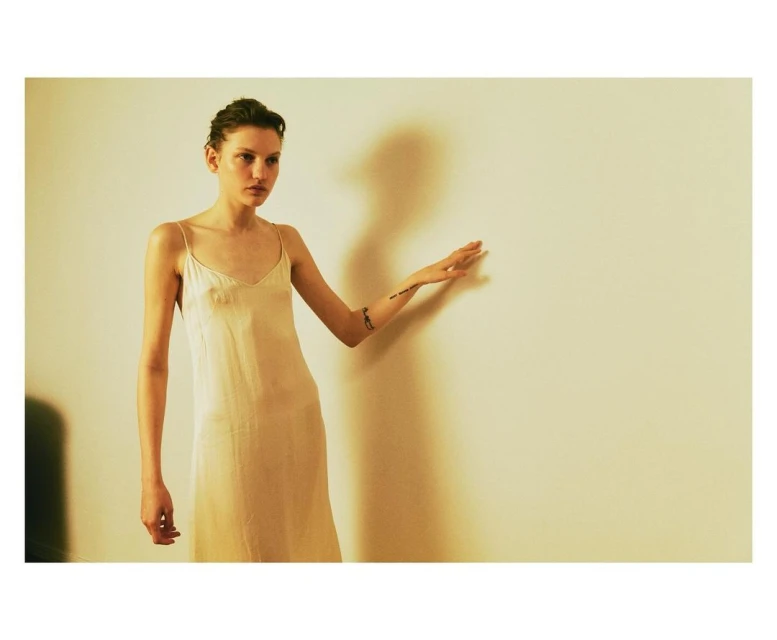 Woman in slip dress stands in front of cream wall with silhouette. Studio lighting accentuates minimalistic fashion and simple beauty.