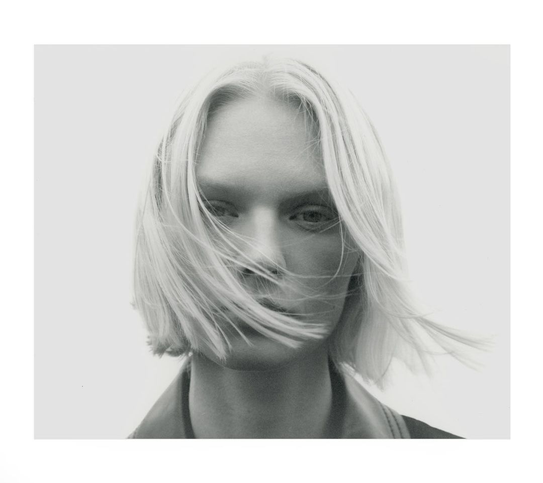 Monochrome portrait of a fair-skinned adult female with platinum blonde hair styled across her face in a light airy studio setting.