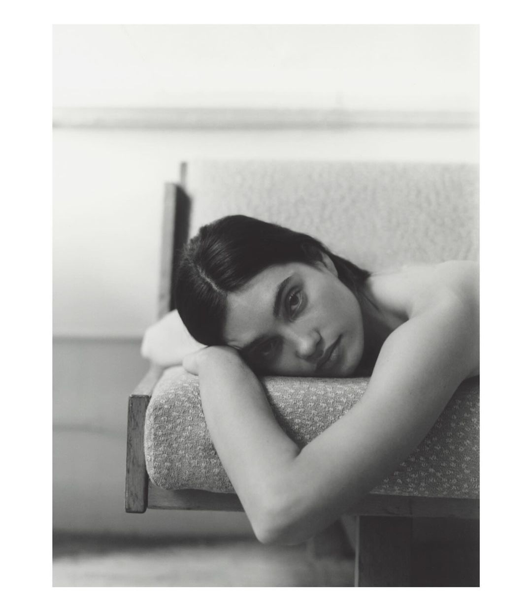Portrait of a woman with dark hair resting on a patterned sofa, looking at the camera with a neutral expression in a monochrome photo.