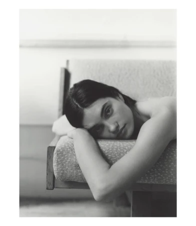 Portrait of a woman with dark hair resting on a patterned sofa, looking at the camera with a neutral expression in a monochrome photo.