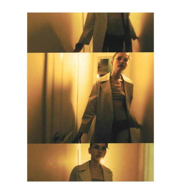 Indoor shot of a young adult woman in a trench coat, standing in a narrow hallway with orange-yellow walls and soft lighting.
