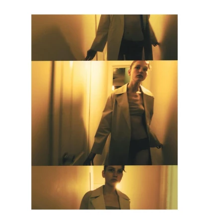 Indoor shot of a young adult woman in a trench coat, standing in a narrow hallway with orange-yellow walls and soft lighting.