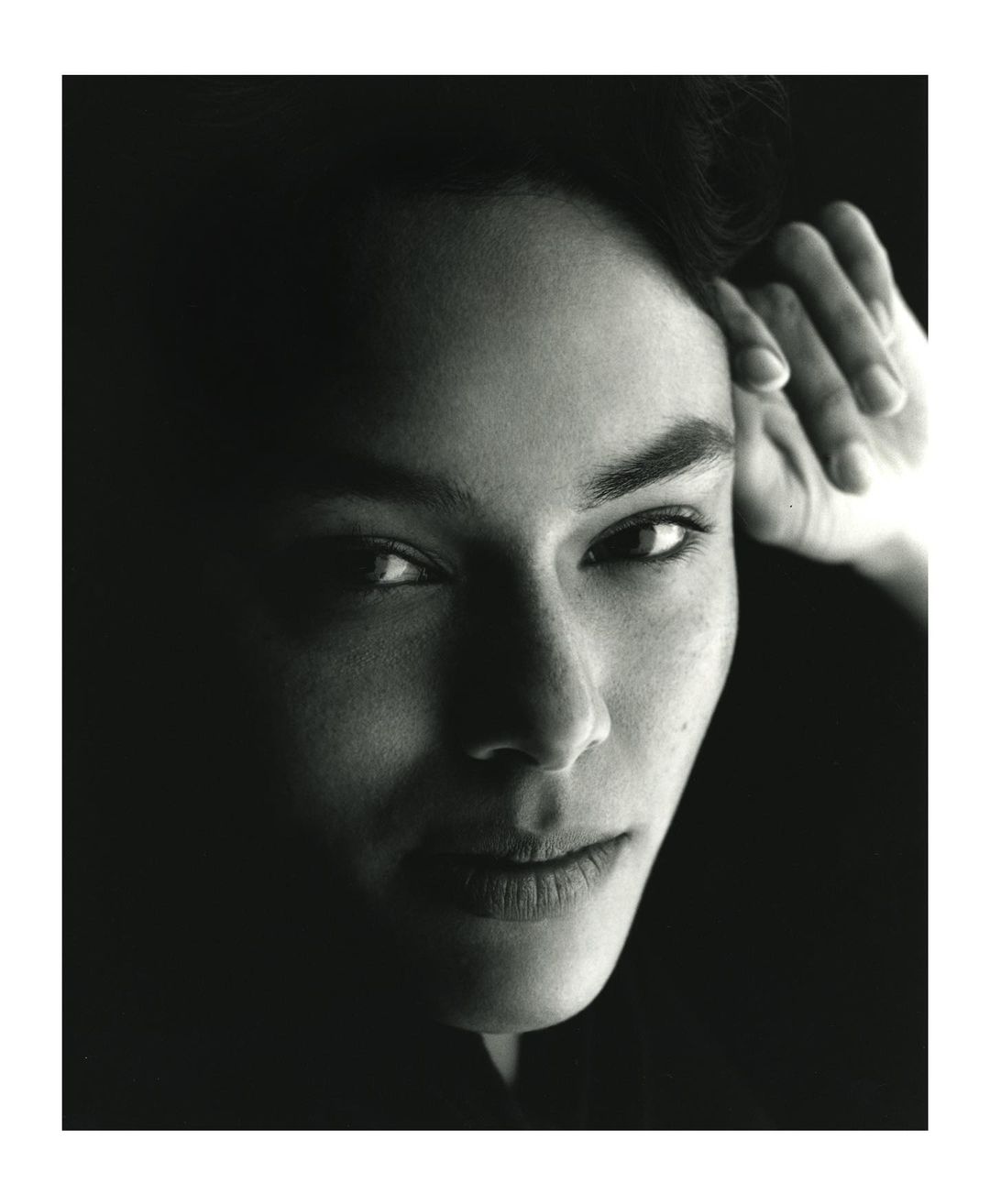 Monochrome portrait of a person looking directly at the camera with their hand near their face against a dark background.