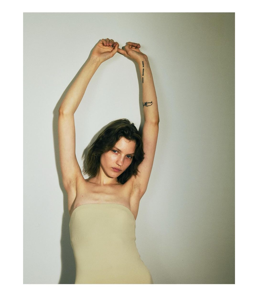 Portrait of a young woman with short wavy hair, wearing a light-colored strapless top, posing with her arms raised against a white wall.
