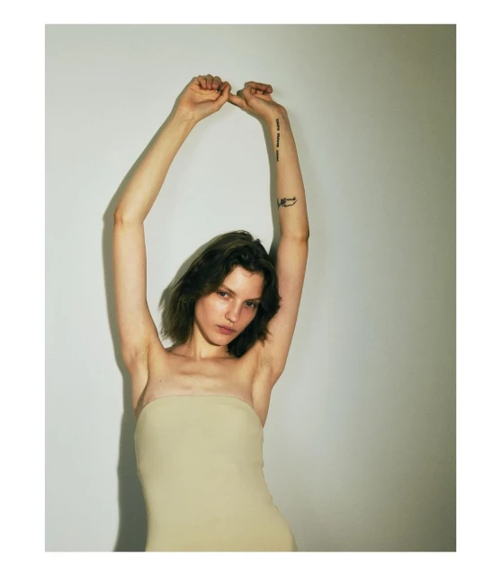 Portrait of a young woman with short wavy hair, wearing a light-colored strapless top, posing with her arms raised against a white wall.