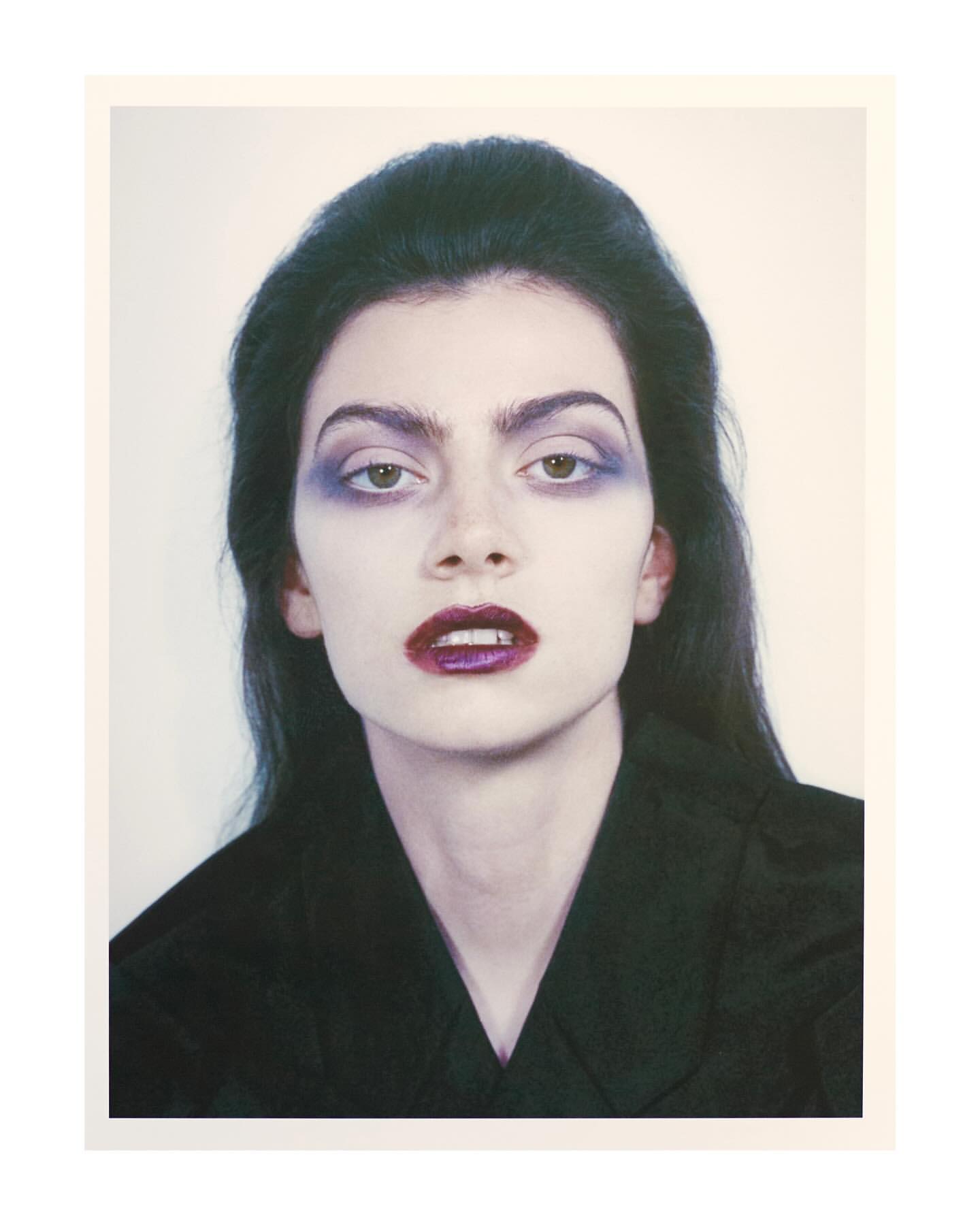 Close-up portrait of a woman with dark hair, wearing purple makeup on her eyes and lips, and a dark v-neck top against a light background.