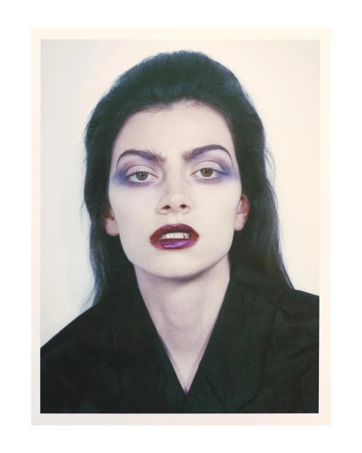 Close-up portrait of a woman with dark hair, wearing purple makeup on her eyes and lips, and a dark v-neck top against a light background.