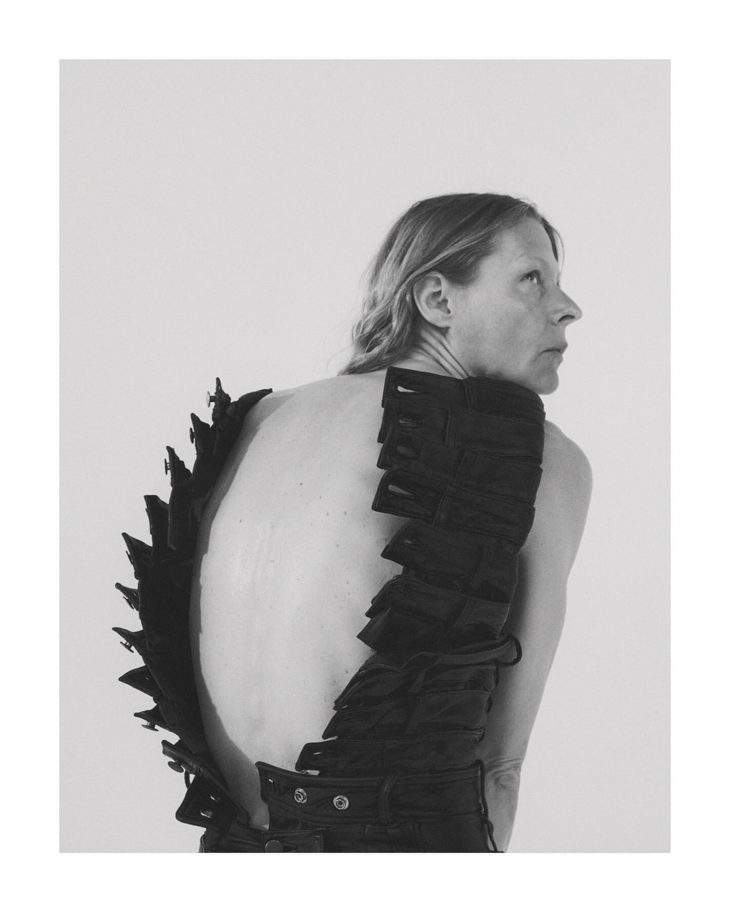 Monochrome portrait of an adult woman with bare back wearing a black textured garment and looking sideways in a bright photo studio.