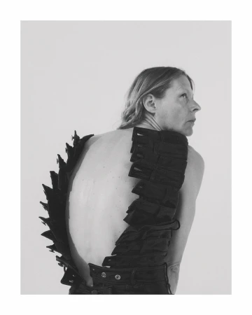 Monochrome portrait of an adult woman with bare back wearing a black textured garment and looking sideways in a bright photo studio.