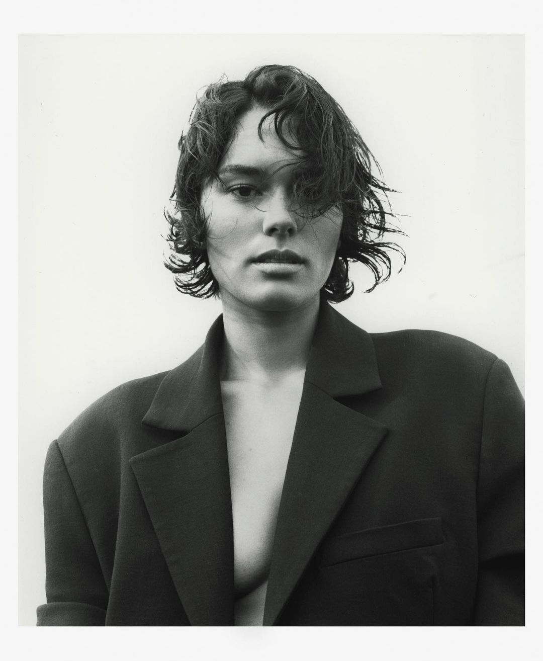 Black-and-white portrait of a person with short, wavy hair and an open blazer, set against a bright white background.