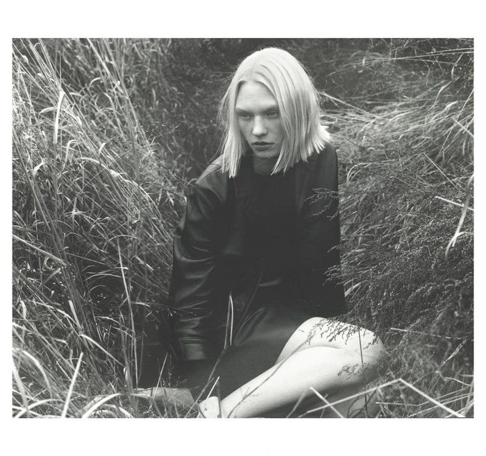 Model with blonde bob hair wearing a dark jacket seated amidst tall overgrown grass in a black and white portrait photograph.