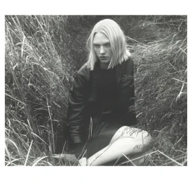 Model with blonde bob hair wearing a dark jacket seated amidst tall overgrown grass in a black and white portrait photograph.