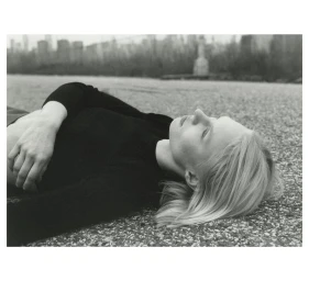 Monochrome portrait of a woman lying on textured ground with a blurred city skyline visible in the distant background.
