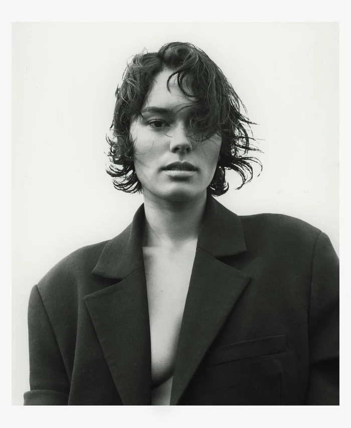 Portrait of a woman with dark wavy hair and a structured black blazer against a plain white background by Andy Harrington
