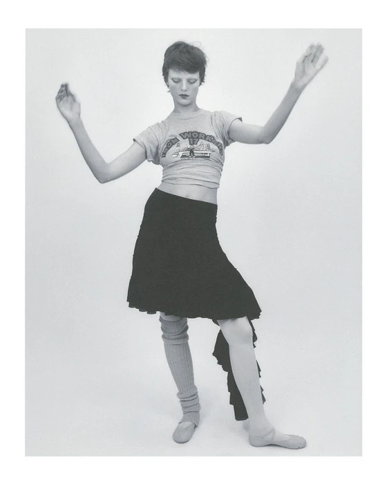 Model with short hair wearing a graphic t-shirt, dark skirt, and leg warmers posing against a plain background