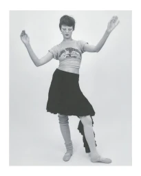 Model with short hair wearing a graphic t-shirt, dark skirt, and leg warmers posing against a plain background