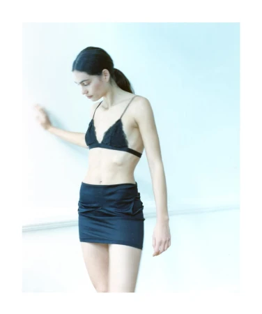 Woman in a black lace bralette and mini skirt posing against a pale wall, photographed by Andy Harrington in Paris.