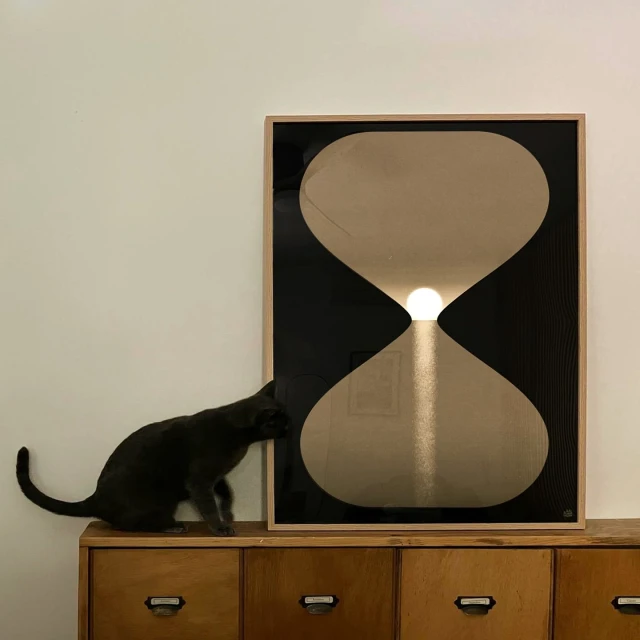 A black cat sniffs a framed, light-brown and black hourglass art print on a wooden cabinet against an off-white wall indoors.