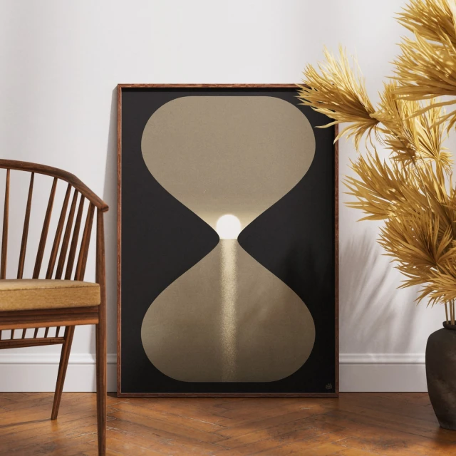 Hourglass-shaped art print in a dark frame displayed in a room with a wood chair and golden dried floral arrangement.