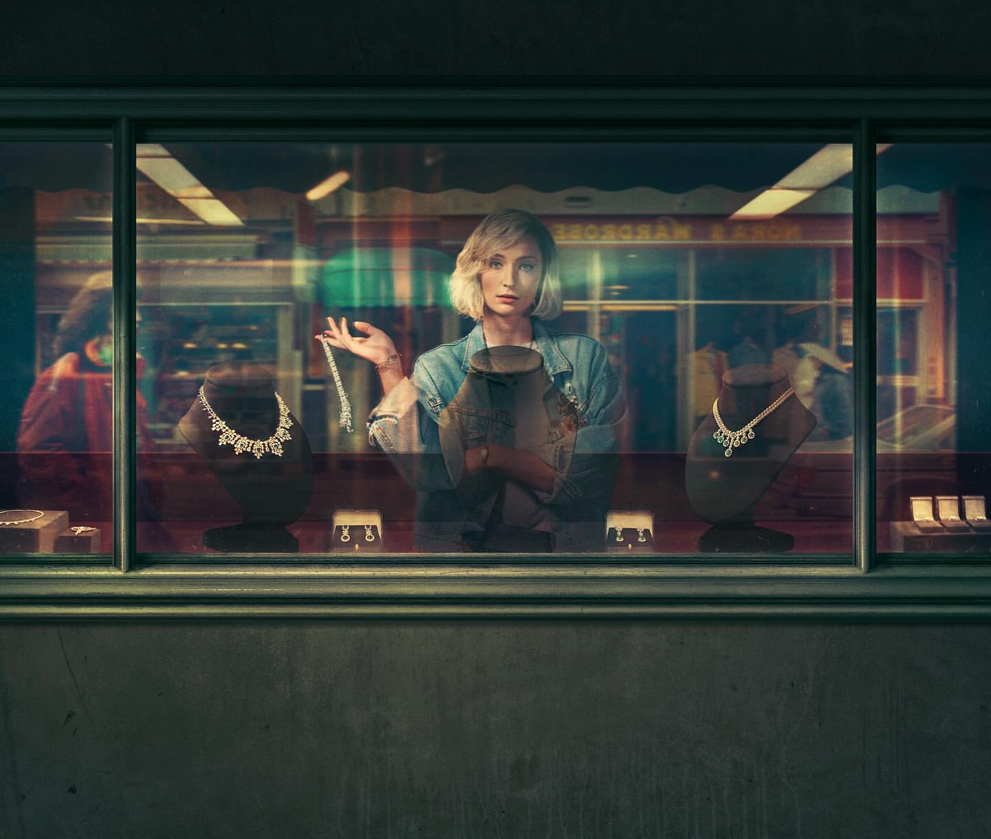 Woman in denim jacket stands in front of a jewelry store window displaying necklaces and earrings in a dimly lit urban setting.