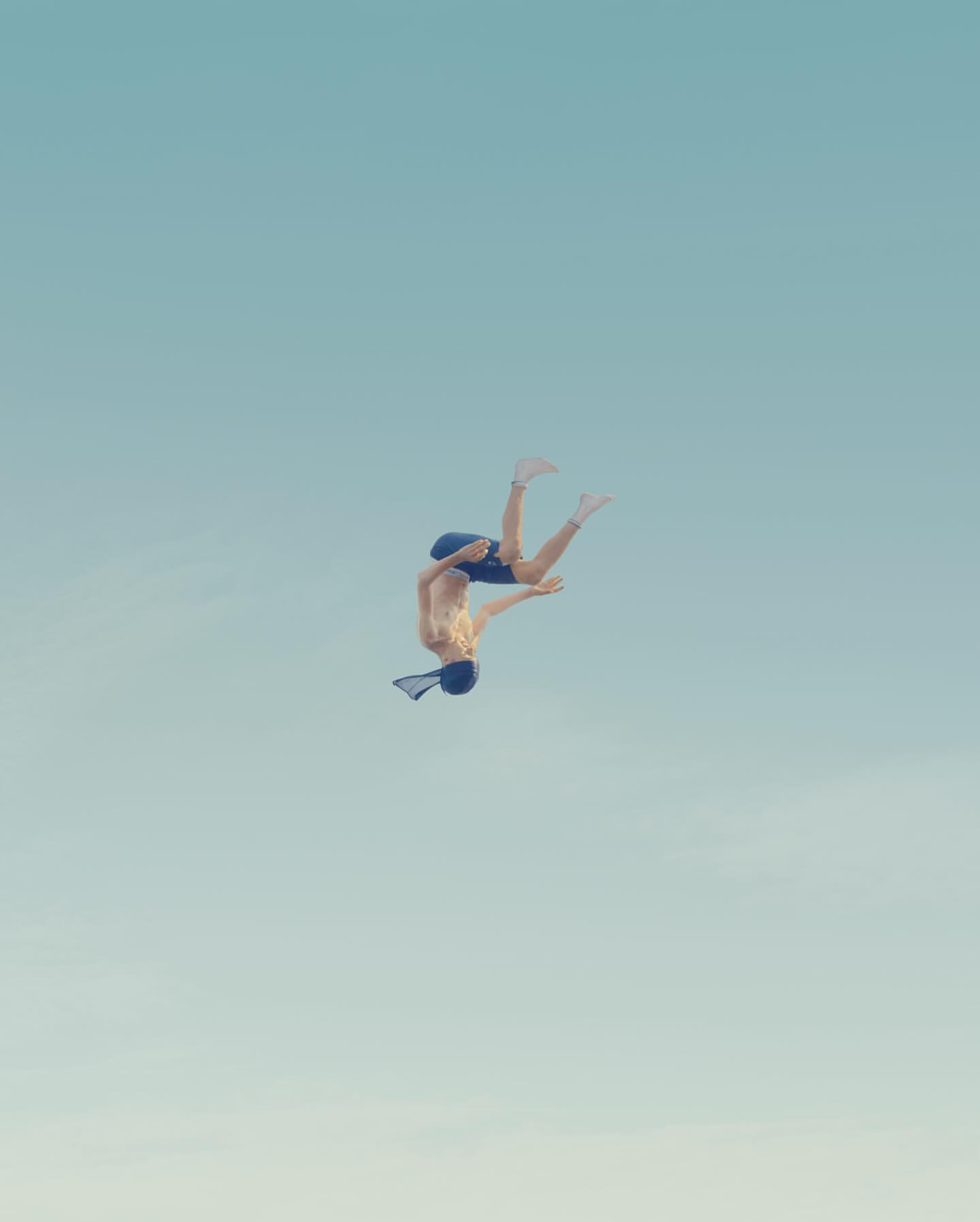 A fair-skinned male athlete performs a backflip against a clear blue sky. The human is wearing dark shorts and a cap. Gymnastics stunt.
