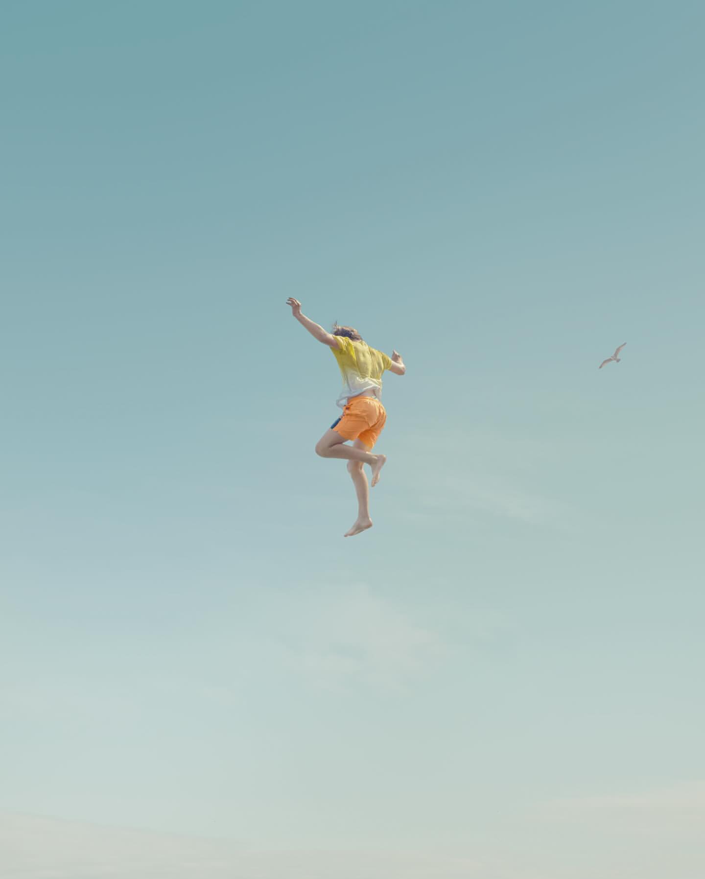 Person jumps in the air against a clear blue sky, wearing orange shorts and a yellow shirt, arms raised and a bird flying in the distance.