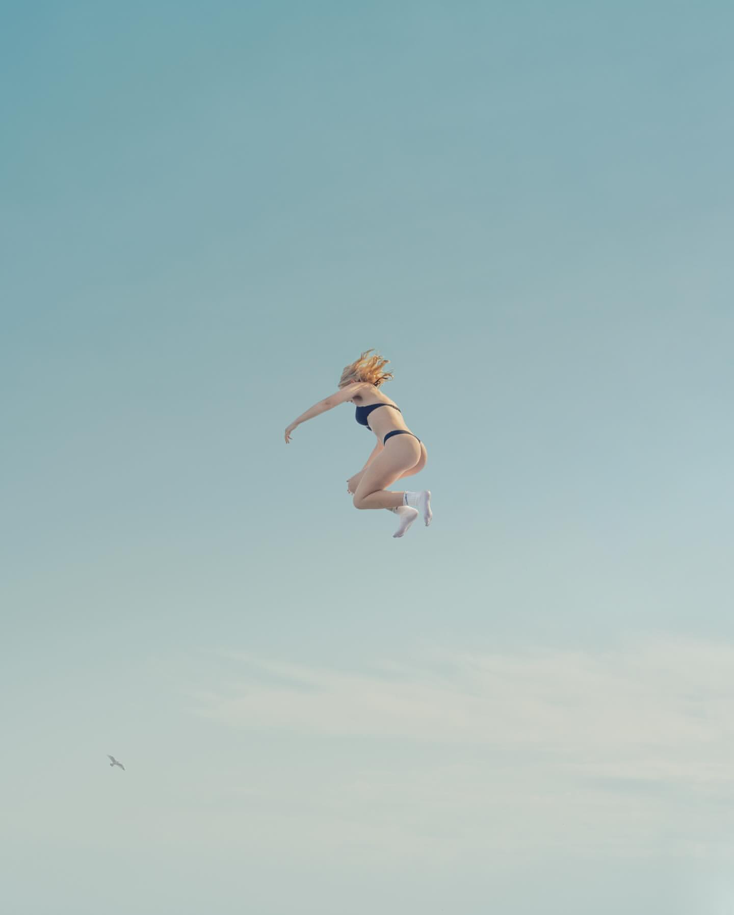 Woman in black bikini mid-air jump against light-blue sky backdrop, creating a sense of freedom and athleticism in the outdoors.
