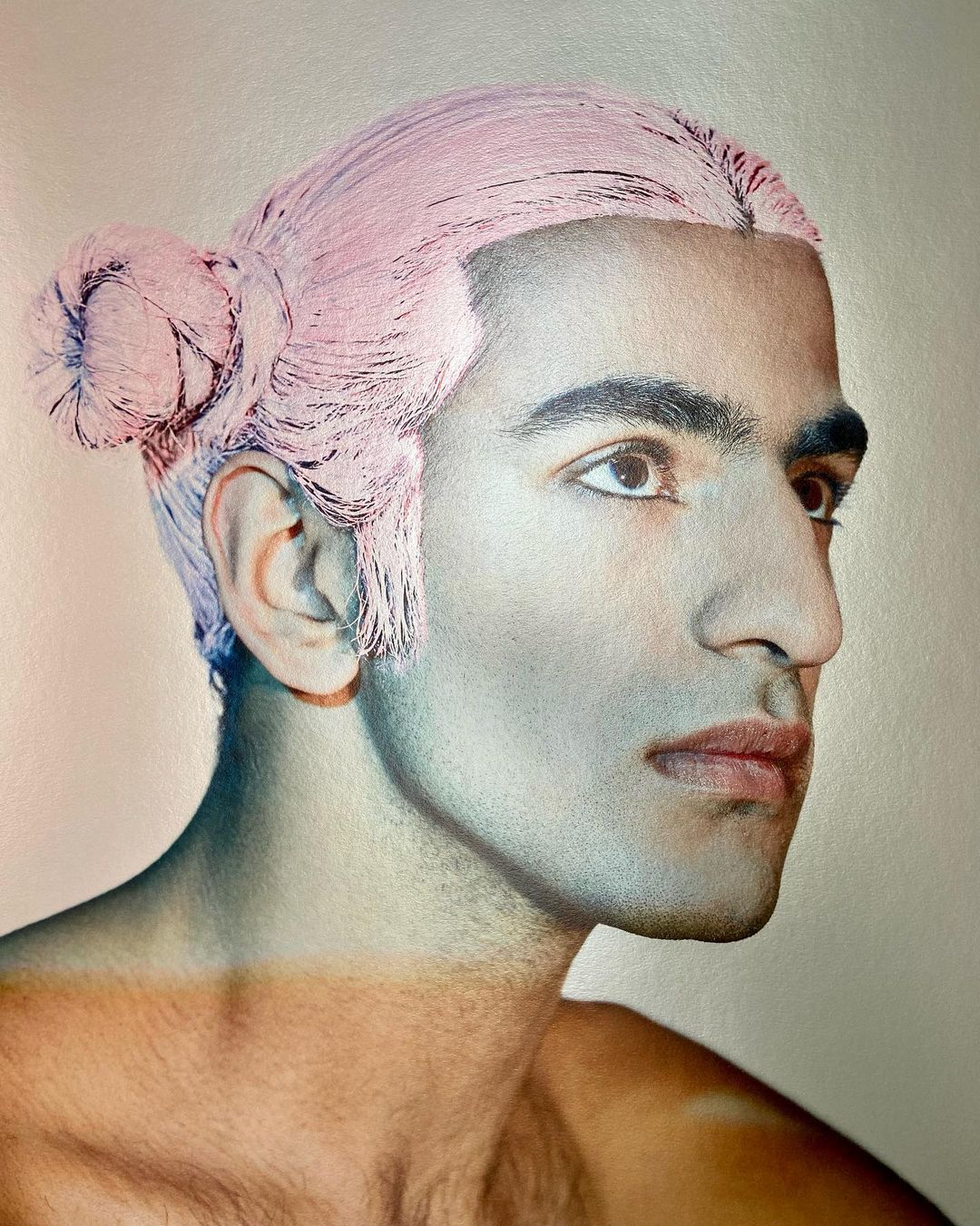 Close-up portrait of a man with pink hair styled in a bun, looking to the right in a studio setting with soft, diffused lighting.