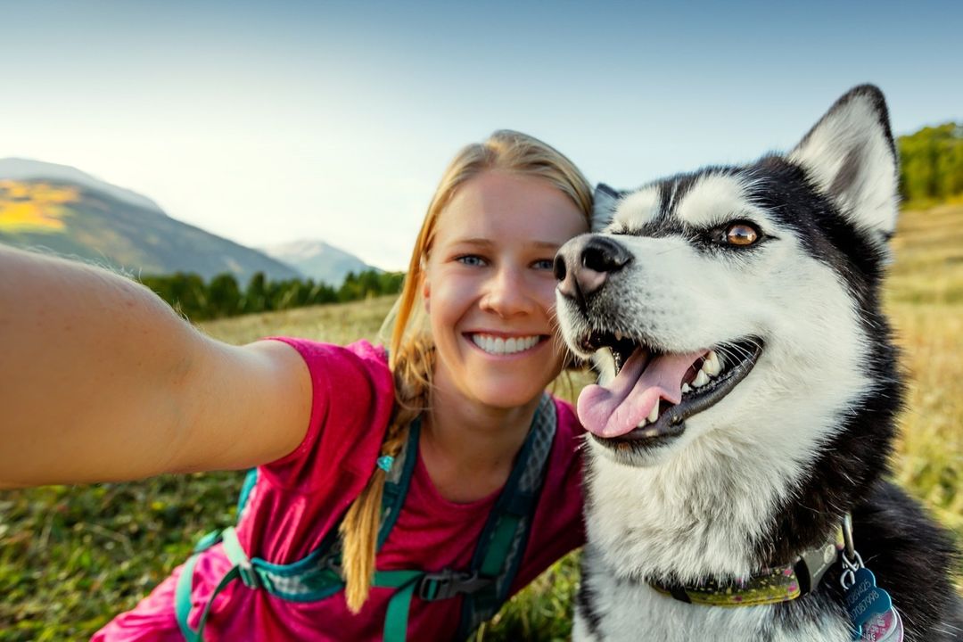 Smiling woman takes a selfie with her husky dog outdoors. Mountains and blue sky are in the background on a sunny day.