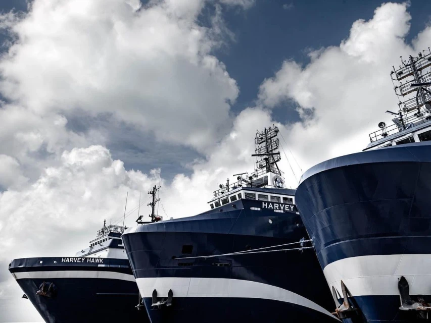 Three navy blue and white offshore supply vessels docked in a harbor, seen from a low angle against a partly cloudy sky, maritime industry.