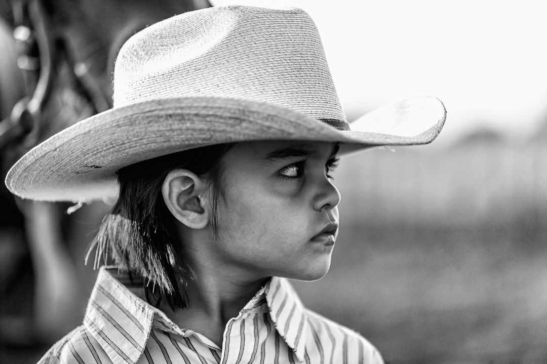 Black and white portrait of child wearing cowboy hat and striped shirt looking off-camera, partial view of horse in background.