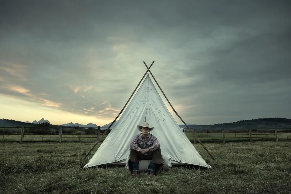 Boy in cowboy hat sits in front of a teepee on a grassy field with a wooden fence, mountains, and a cloudy sky in the background.