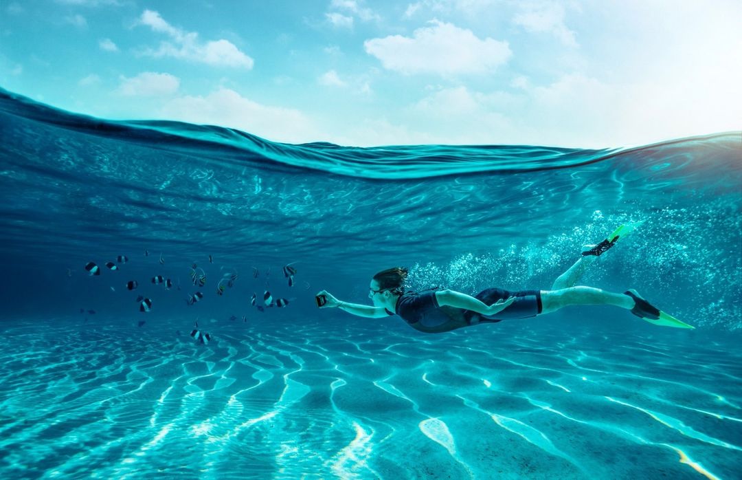 Person swimming underwater with fish in a clear turquoise ocean, wearing flippers and a wetsuit while holding a camera.