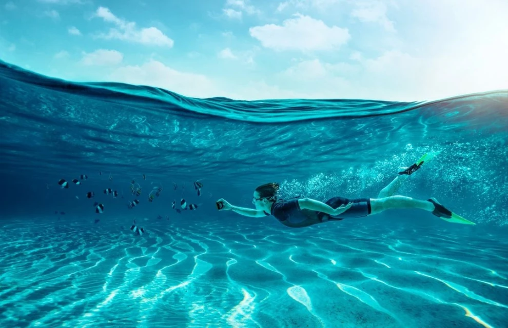 Person swimming underwater with fish in a clear turquoise ocean, wearing flippers and a wetsuit while holding a camera.