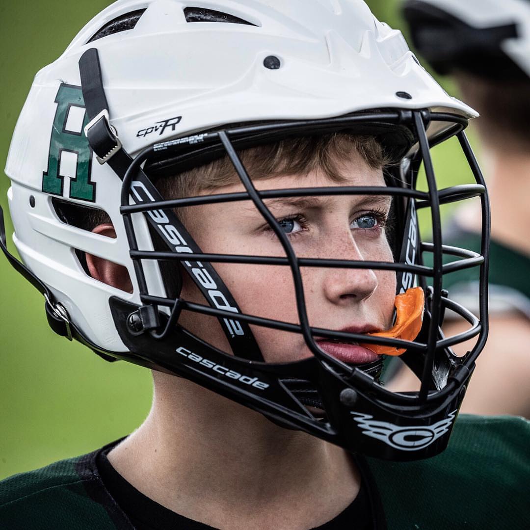 Close-up of a young male athlete wearing a white Cascade lacrosse helmet with a green logo, black facemask, and an orange mouthguard.