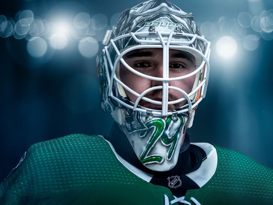 Close-up portrait of a hockey goaltender wearing a helmet, jersey, and pads against a blurred blue background with bokeh.