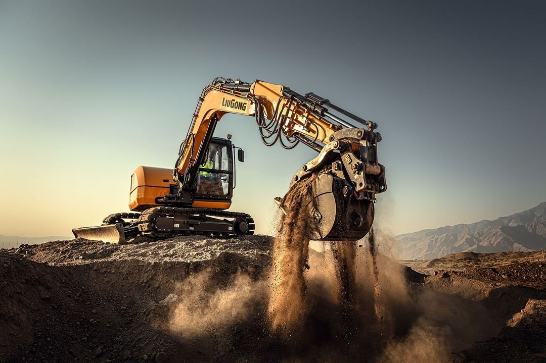 Yellow LiuGong excavator digging in a dirt pile with mountains in the background against a clear sky in an outdoor environment.