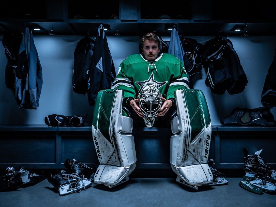 Hockey goalie in uniform sits in locker room holding his helmet. Pads and skates are visible around him with a serious facial expression.
