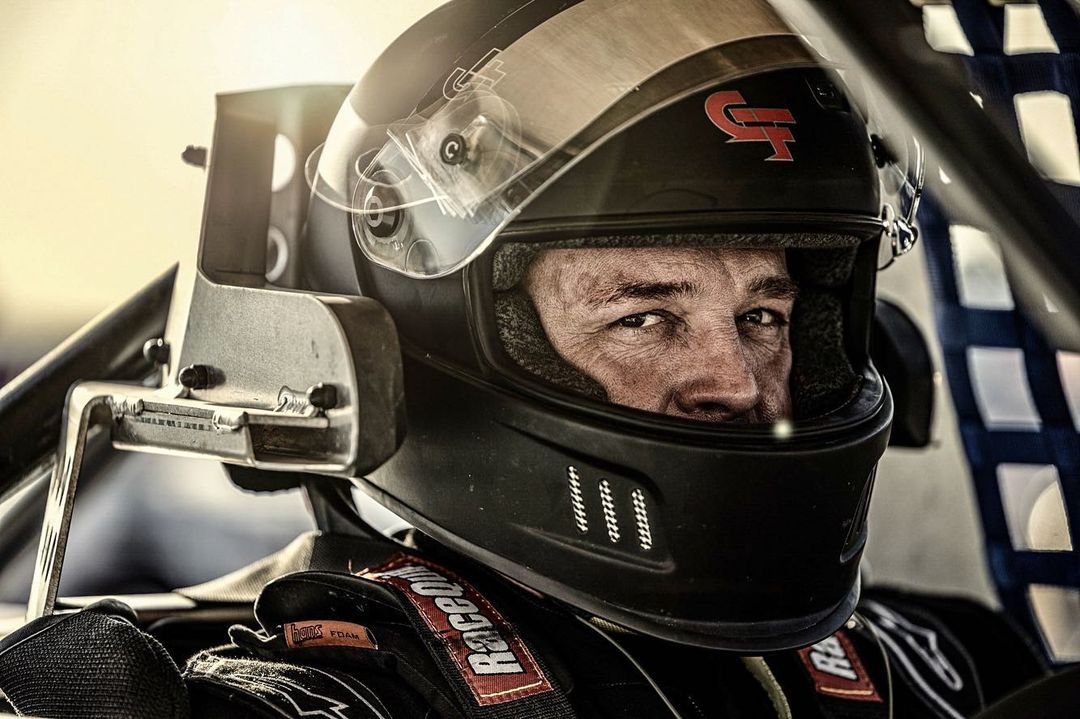 Close-up of a concentrated race car driver in a black helmet, captured indoors with dramatic lighting and high contrast.