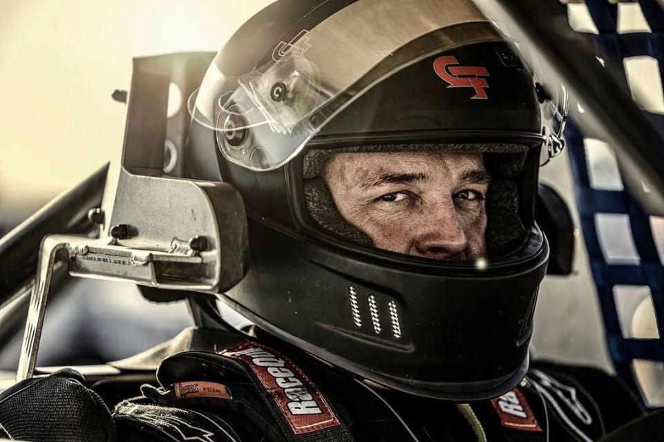 Close-up of a concentrated race car driver in a black helmet, captured indoors with dramatic lighting and high contrast.