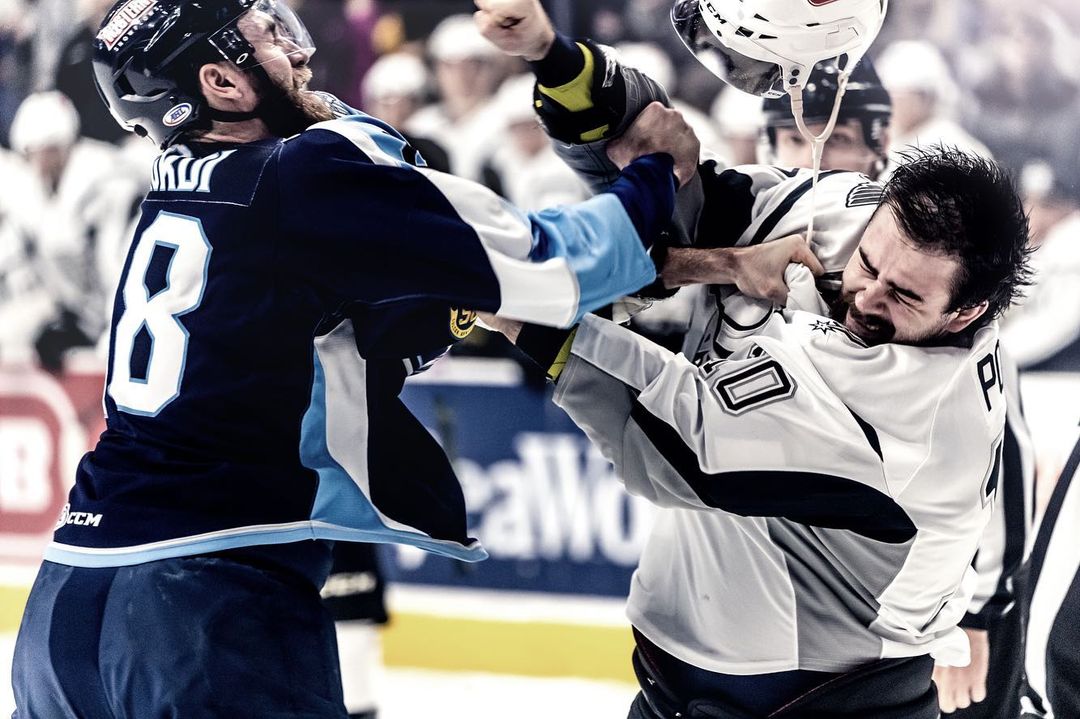 Two adult male ice hockey players fight during a game in a stadium, one wearing white and the other wearing navy blue and light blue.