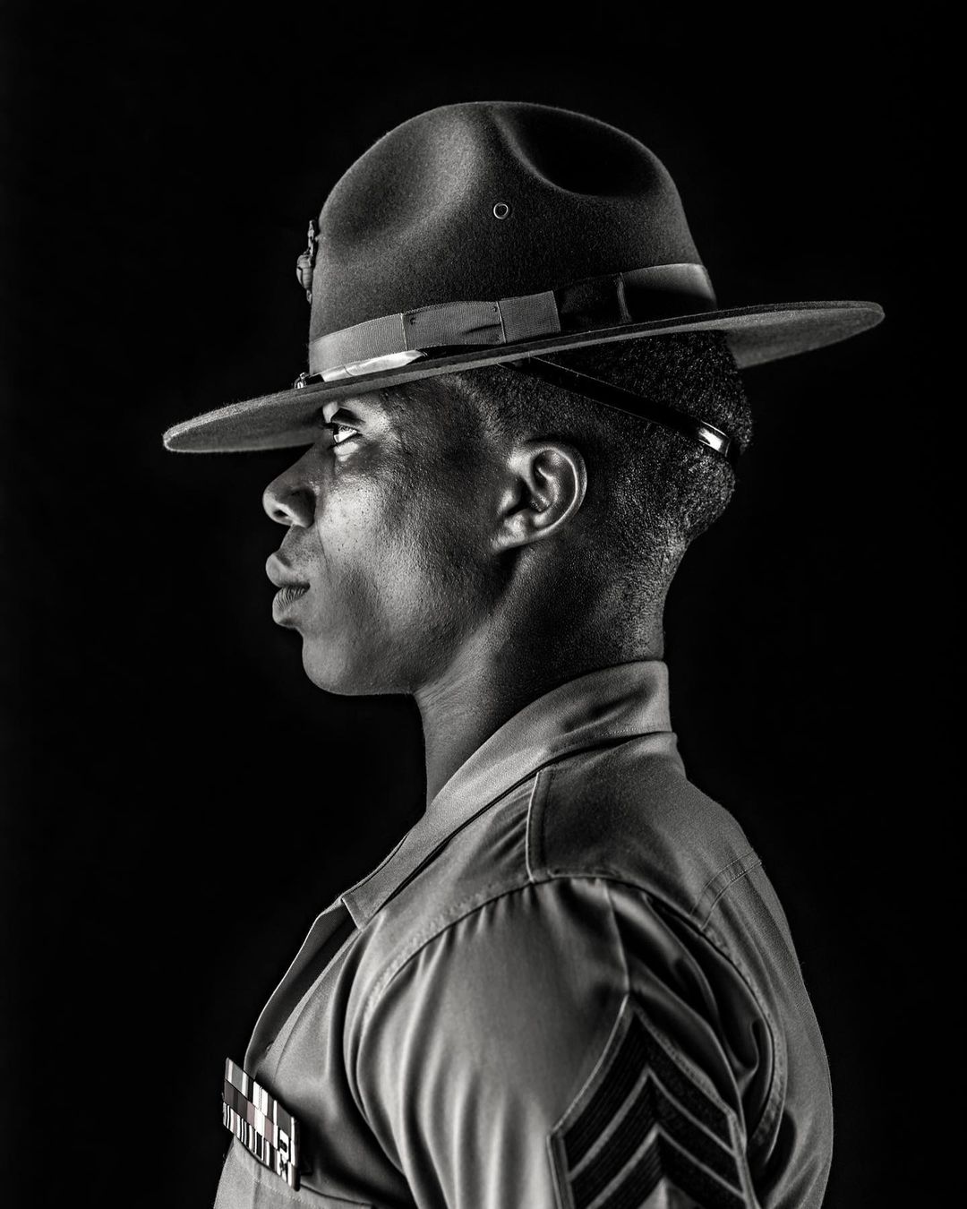 Profile portrait of a serious African-American military man wearing a formal uniform and a drill sergeant hat against a black background.