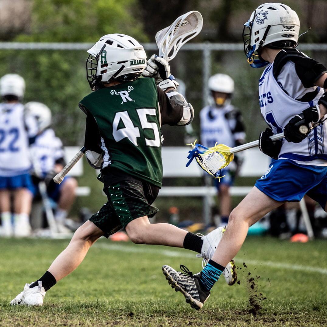 Two young lacrosse players competing on a grassy field, one in green uniform and the other in blue and white, outdoors.
