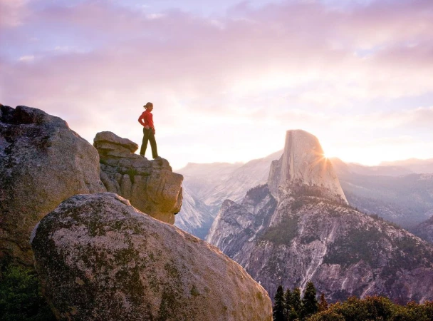 Woman stands on a rocky cliff, overlooking a mountainous landscape with Half Dome in the background, silhouetted against a pastel sky.