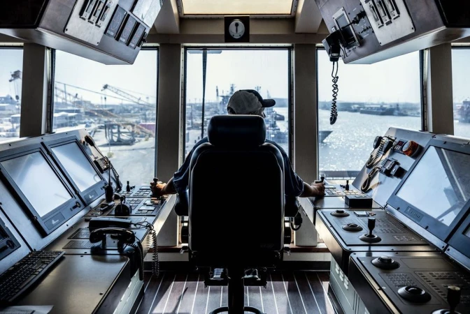 Man operates a vessel in an industrial port, using ship controls inside a control room, with a harbor visible in the background.