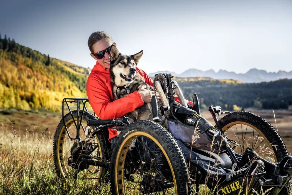 Woman smiles and hugs dog in adaptive bike outdoors, sunny, mountains in background, adventure, travel, sport, active lifestyle.