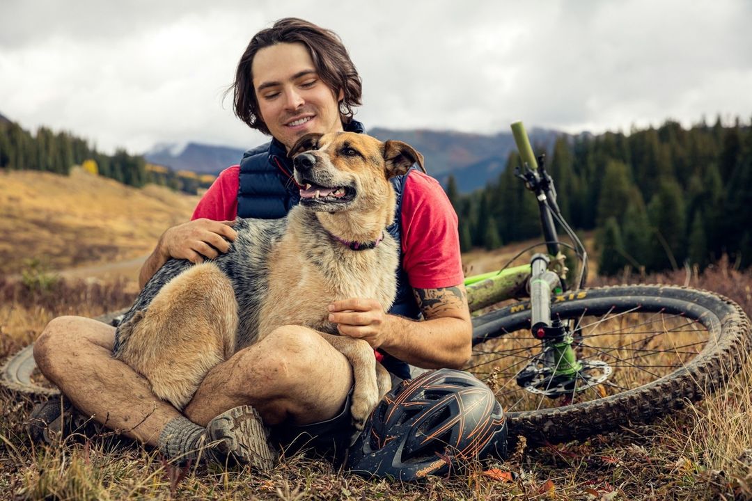 Young man smiles, embracing dog outdoors near mountain bike and helmet on a hillside with fall foliage and distant mountains.